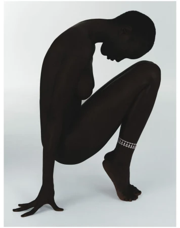 Artistic nude studio shot of a dark-skinned adult female in a crouched pose with head down and an anklet on her left ankle against a white background.