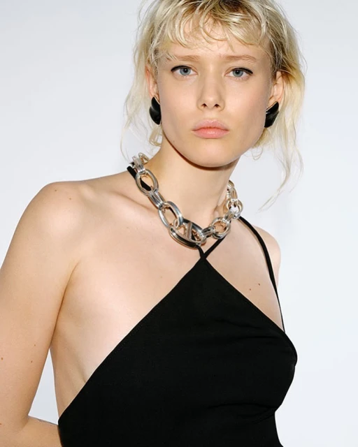Close-up of a young adult female model with pale skin and blonde hair wearing a black dress and a silver chain necklace in a studio.