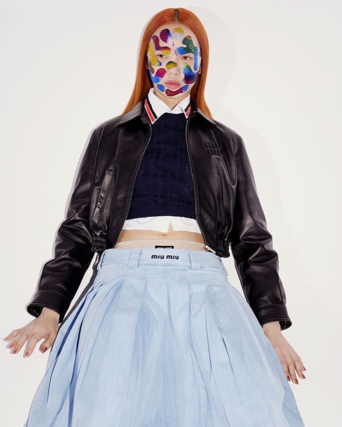 Fashion editorial featuring a woman with red hair and vibrant face paint, wearing a black leather jacket and a light blue Miu Miu skirt in a studio.