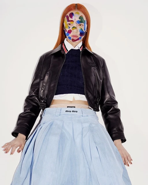 Fashion editorial featuring a woman with red hair and vibrant face paint, wearing a black leather jacket and a light blue Miu Miu skirt in a studio.