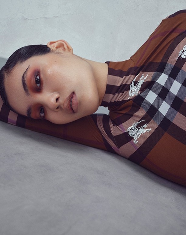 Portrait of a woman laying down wearing a brown Burberry plaid turtleneck. She has smokey makeup and slicked back hair against a grey background.