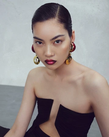 Close-up portrait of an East Asian woman with slicked-back hair, dark red lipstick, and large gold earrings wearing a strapless black top.