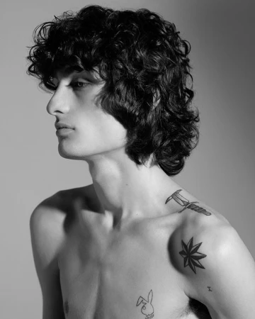 Monochrome portrait of a young man with curly dark hair and tattoos in profile against a plain grey background