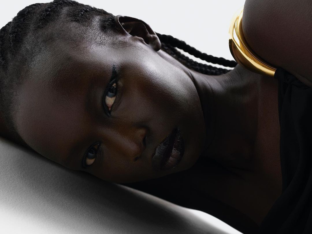 Close-up portrait of a Black woman with braided hair wearing a thick gold cuff bracelet and black clothing against a white backdrop.