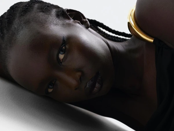 Close-up portrait of a Black woman with braided hair wearing a thick gold cuff bracelet and black clothing against a white backdrop.