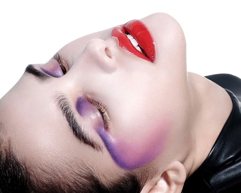 Close-up beauty shot of a woman with vibrant purple eyeshadow and bold red lipstick against a stark white background.