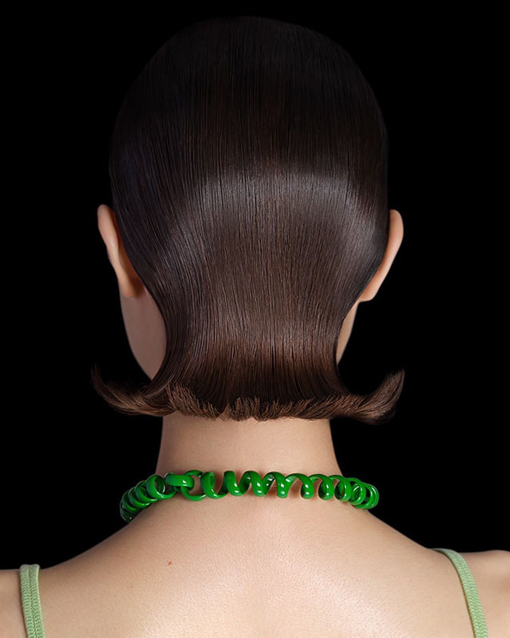 Back view of woman with short, glossy dark brown hair and a bright green spiral necklace against a stark black background.