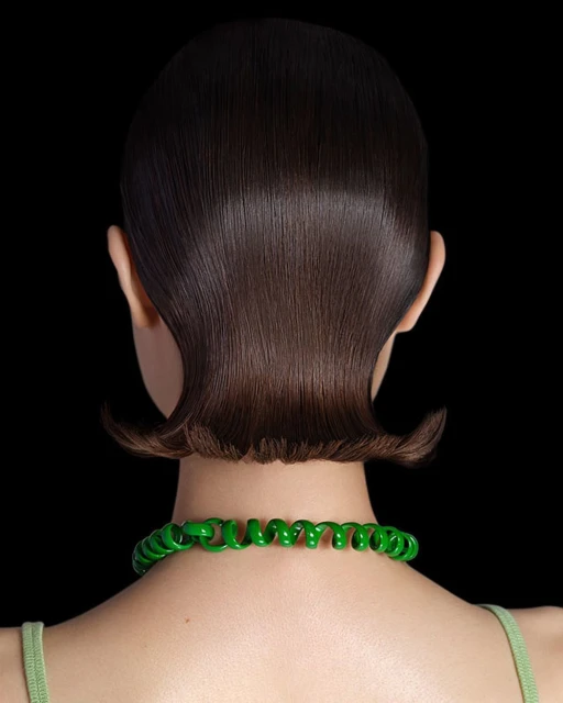 Back view of woman with short, glossy dark brown hair and a bright green spiral necklace against a stark black background.