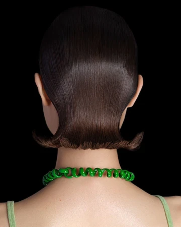 Back view of woman with short, glossy dark brown hair and a bright green spiral necklace against a stark black background.