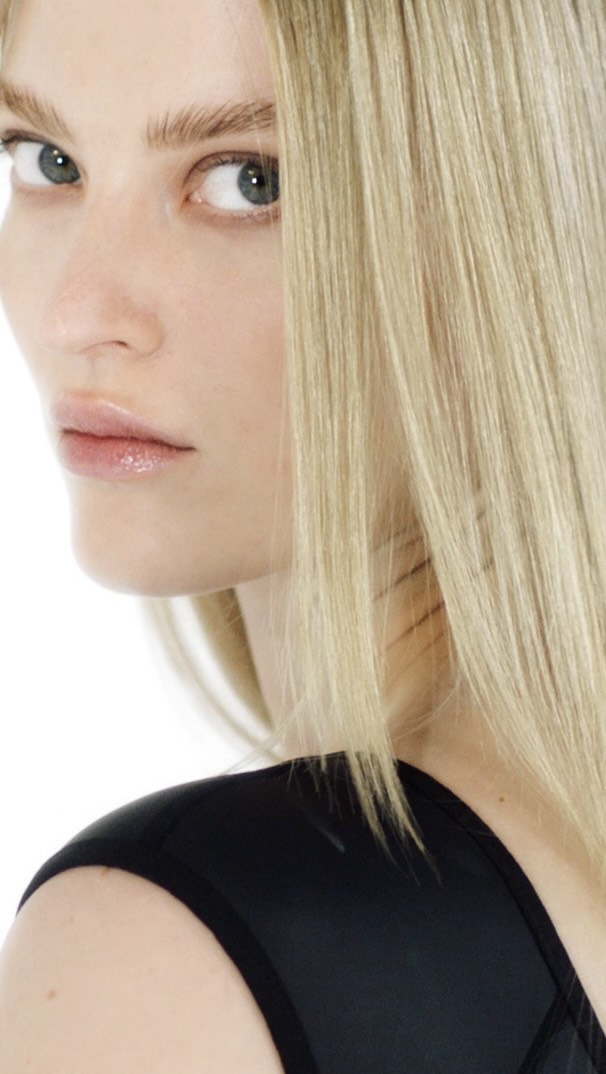 Close-up portrait of a blonde woman in a black top looking over her shoulder against a bright white background.