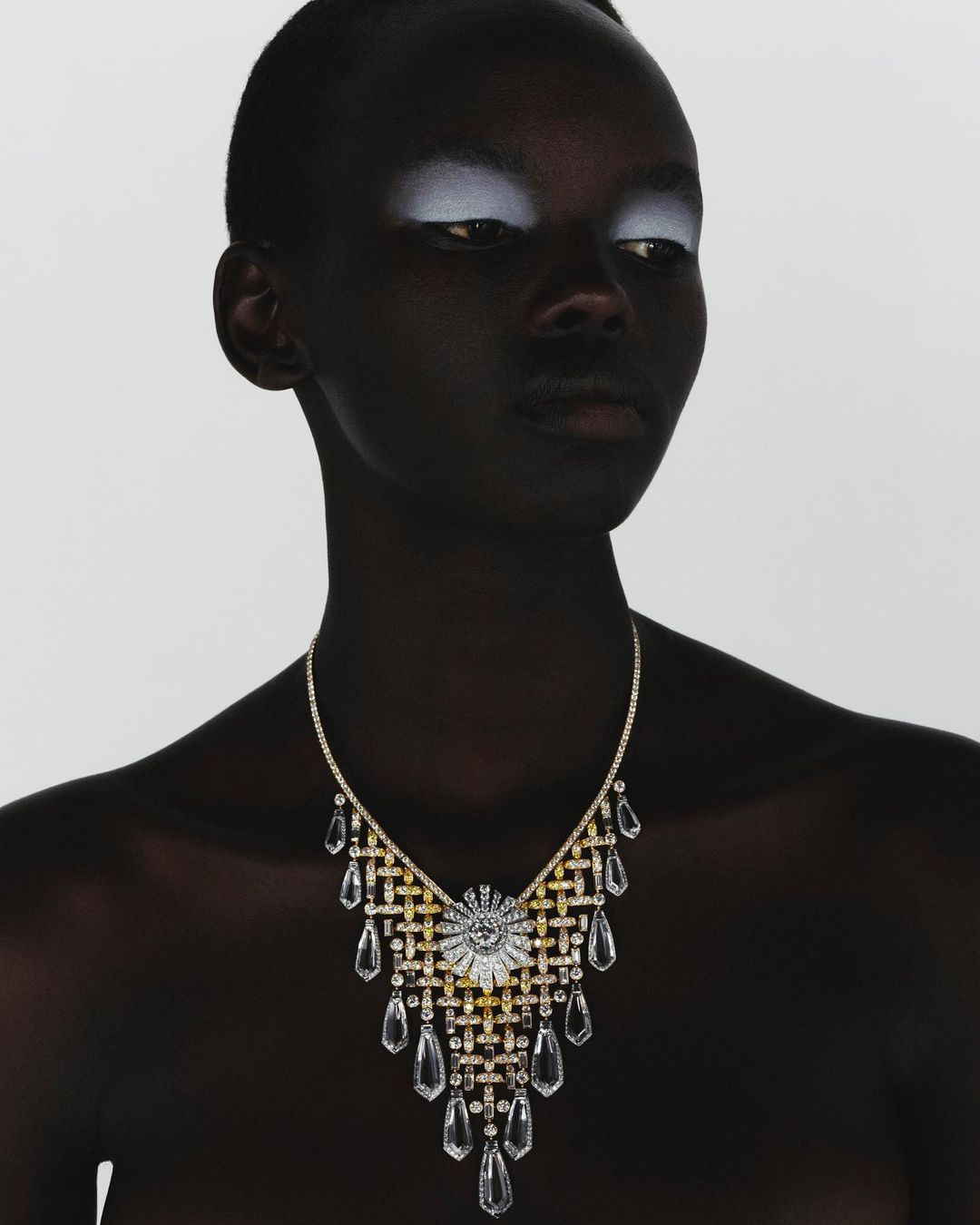 Headshot of a dark-skinned woman with shimmering white eyeshadow wearing a gold and clear crystal statement necklace on a white background.