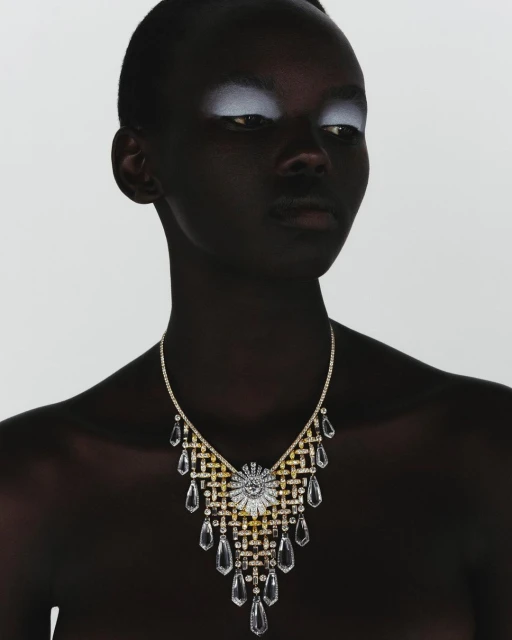 Headshot of a dark-skinned woman with shimmering white eyeshadow wearing a gold and clear crystal statement necklace on a white background.