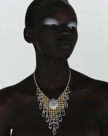 Headshot of a dark-skinned woman with shimmering white eyeshadow wearing a gold and clear crystal statement necklace on a white background.