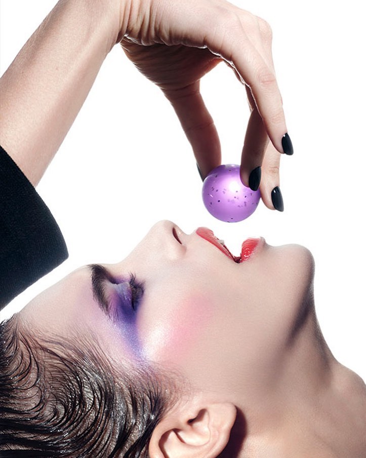 Fashion shot of woman with artistic makeup, purple eye shadow, pale skin, red lipstick, hand holding purple ball, white background.