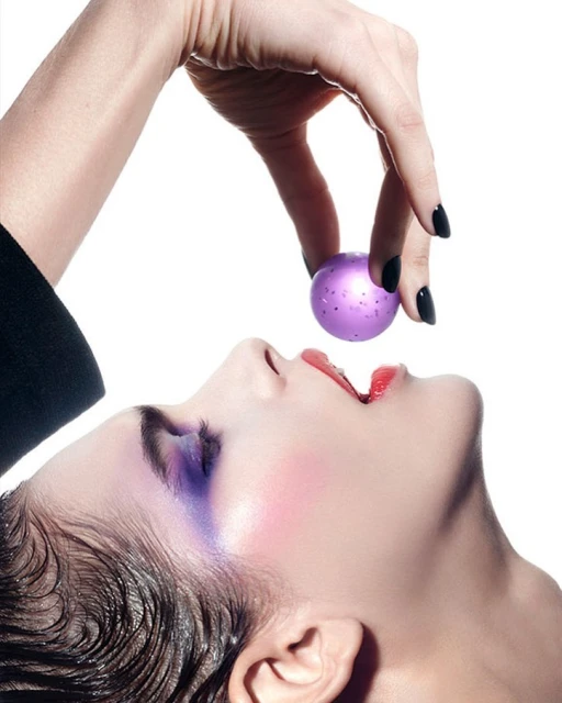 Fashion shot of woman with artistic makeup, purple eye shadow, pale skin, red lipstick, hand holding purple ball, white background.