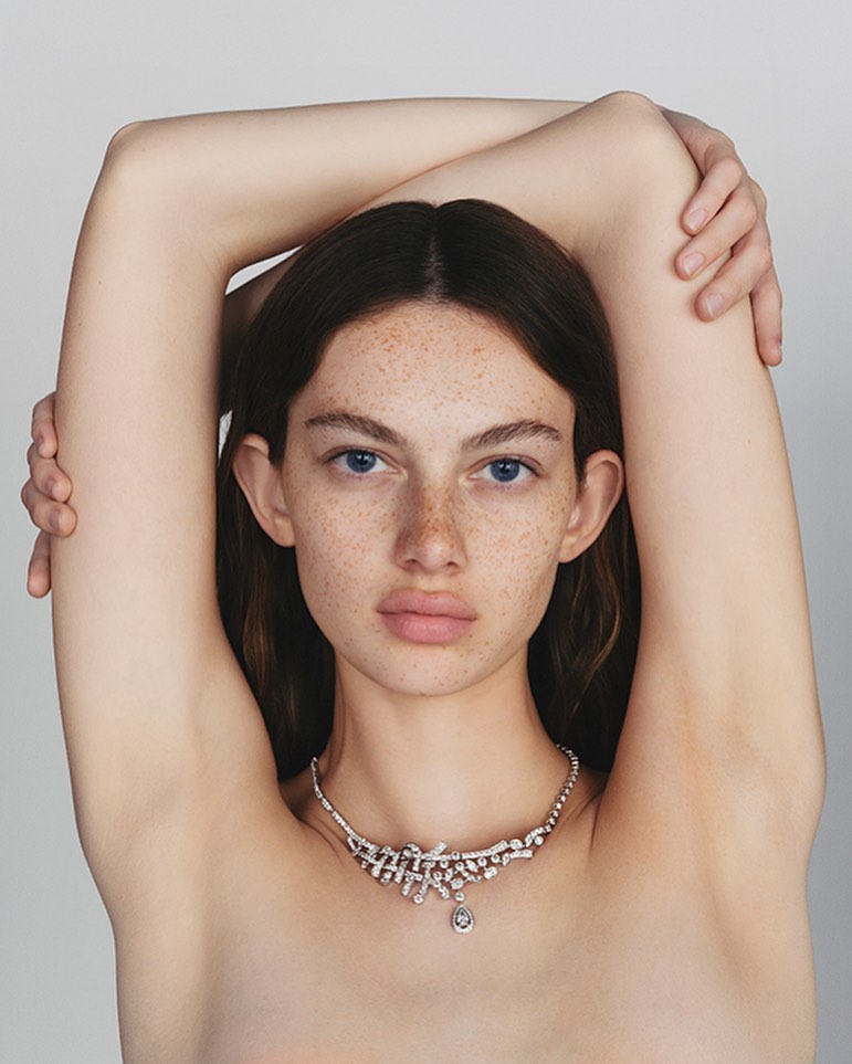 Close-up portrait of a freckled woman with arms raised behind her head, wearing a diamond necklace on white background