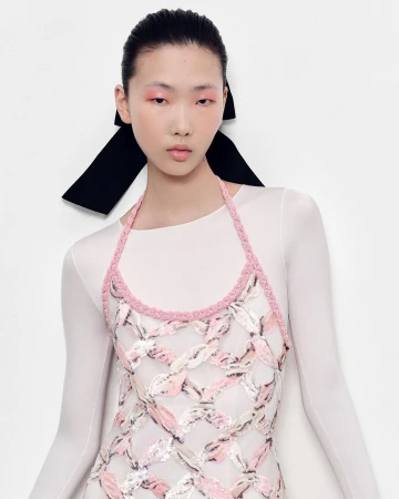 Model with slicked-back hair wearing a pink halter dress and white sheer long-sleeve top against a white backdrop.