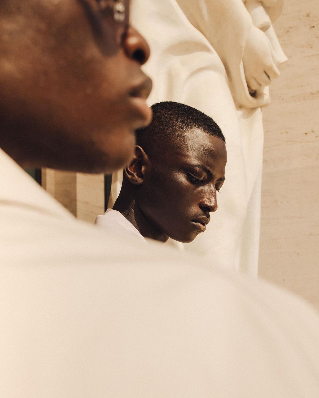 Close-up shot of two African American men with short haircuts, one partially out of frame, against a beige background with a white statue.