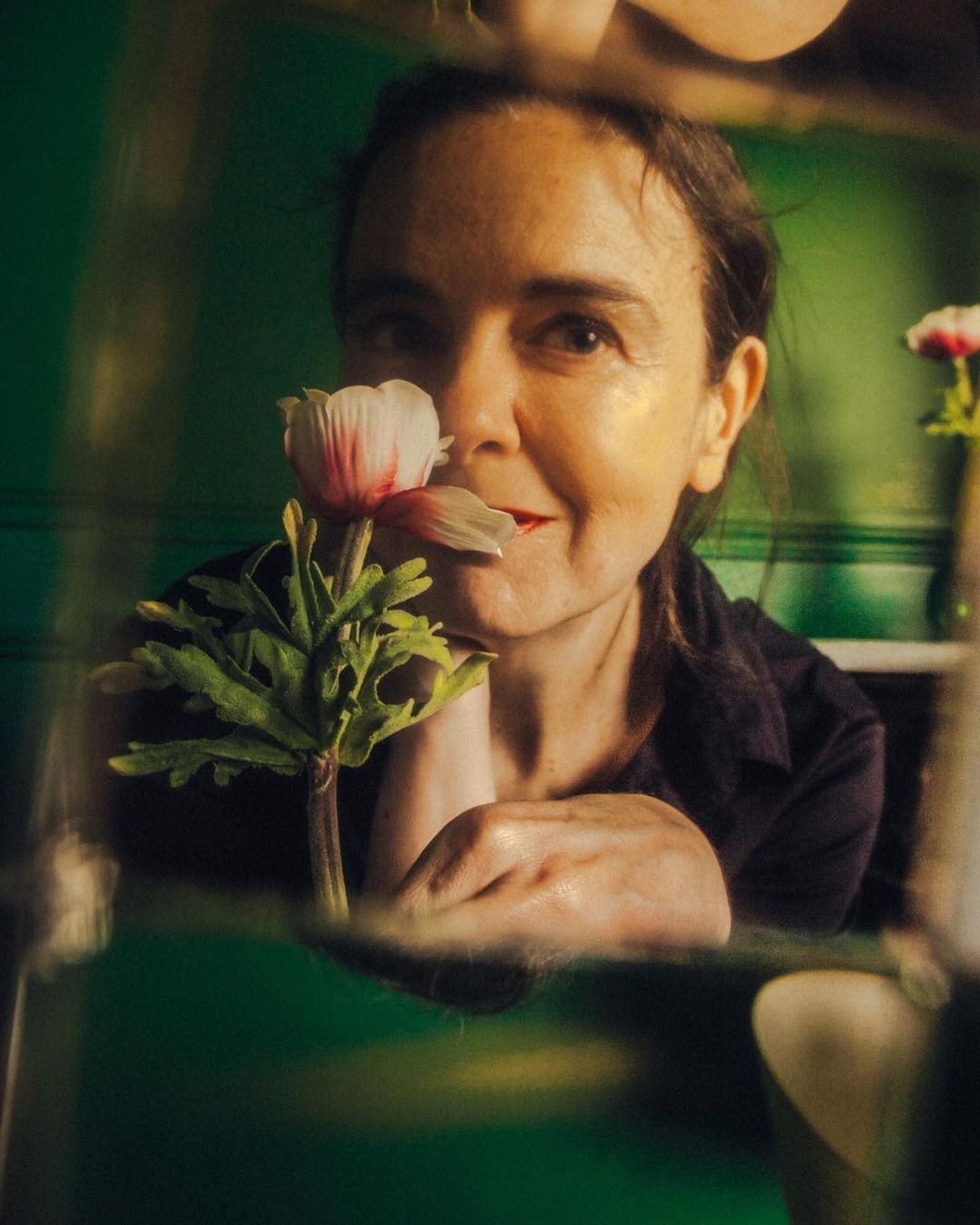 Portrait of a mature woman holding a white and pink flower up to her face in an indoor setting with a blurred green background.