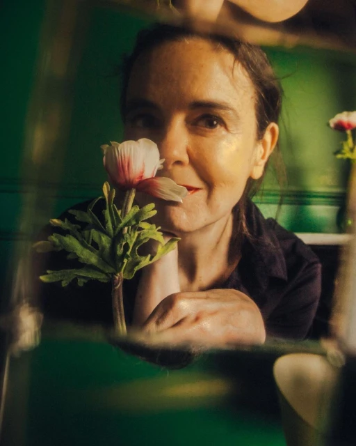 Portrait of a mature woman holding a white and pink flower up to her face in an indoor setting with a blurred green background.