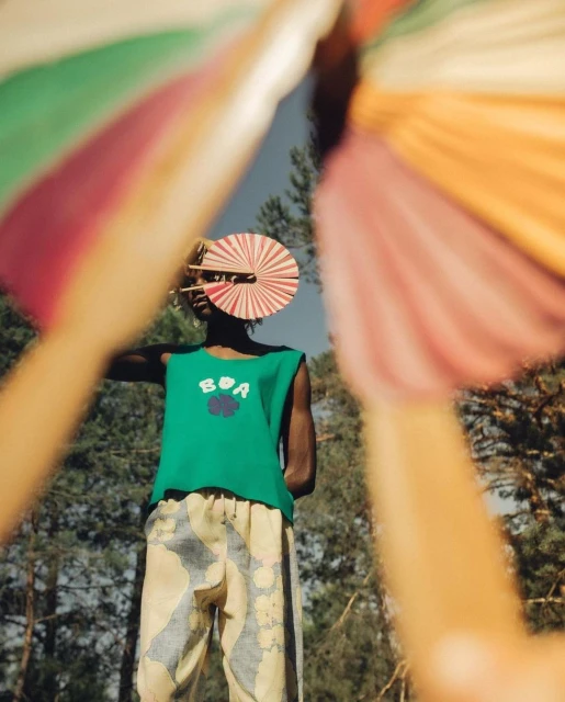 Fashionable person posing outdoors with a hand fan in front of face, wearing a sleeveless shirt and patterned pants, trees in background.