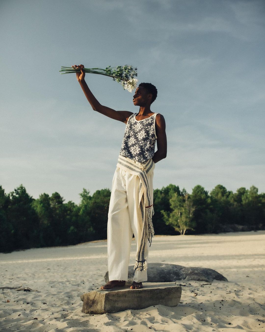 A slender black woman in a patterned tank top and white pants poses with a bouquet of white flowers in a sunny, natural landscape.
