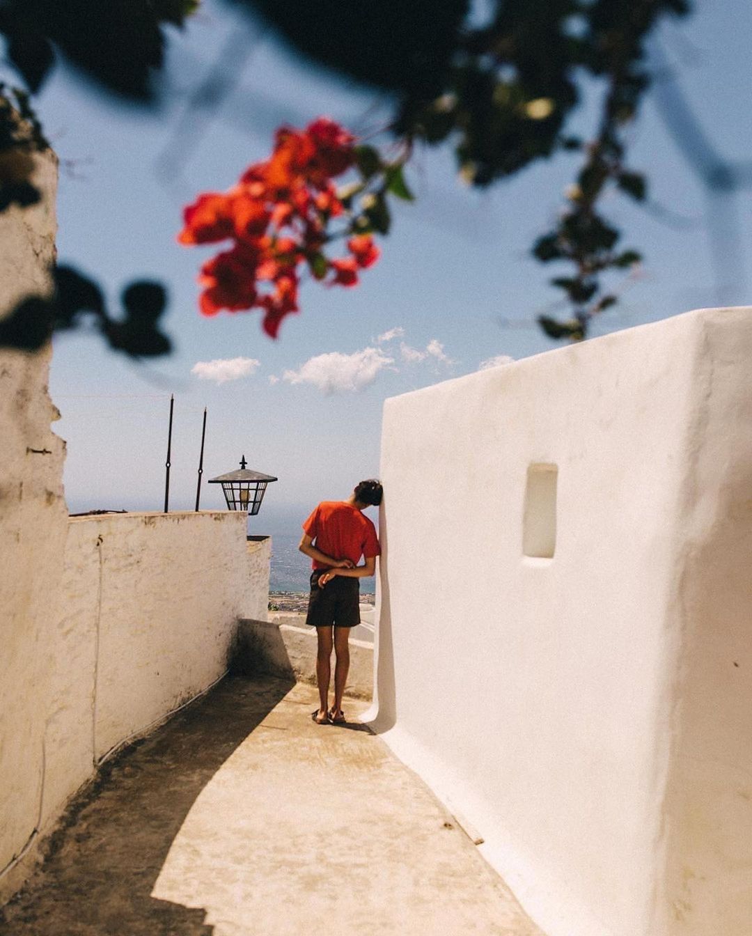 Person in red shirt stands in alleyway with white walls and red flowers in the foreground under a sunny blue sky, Santorini, Greece.