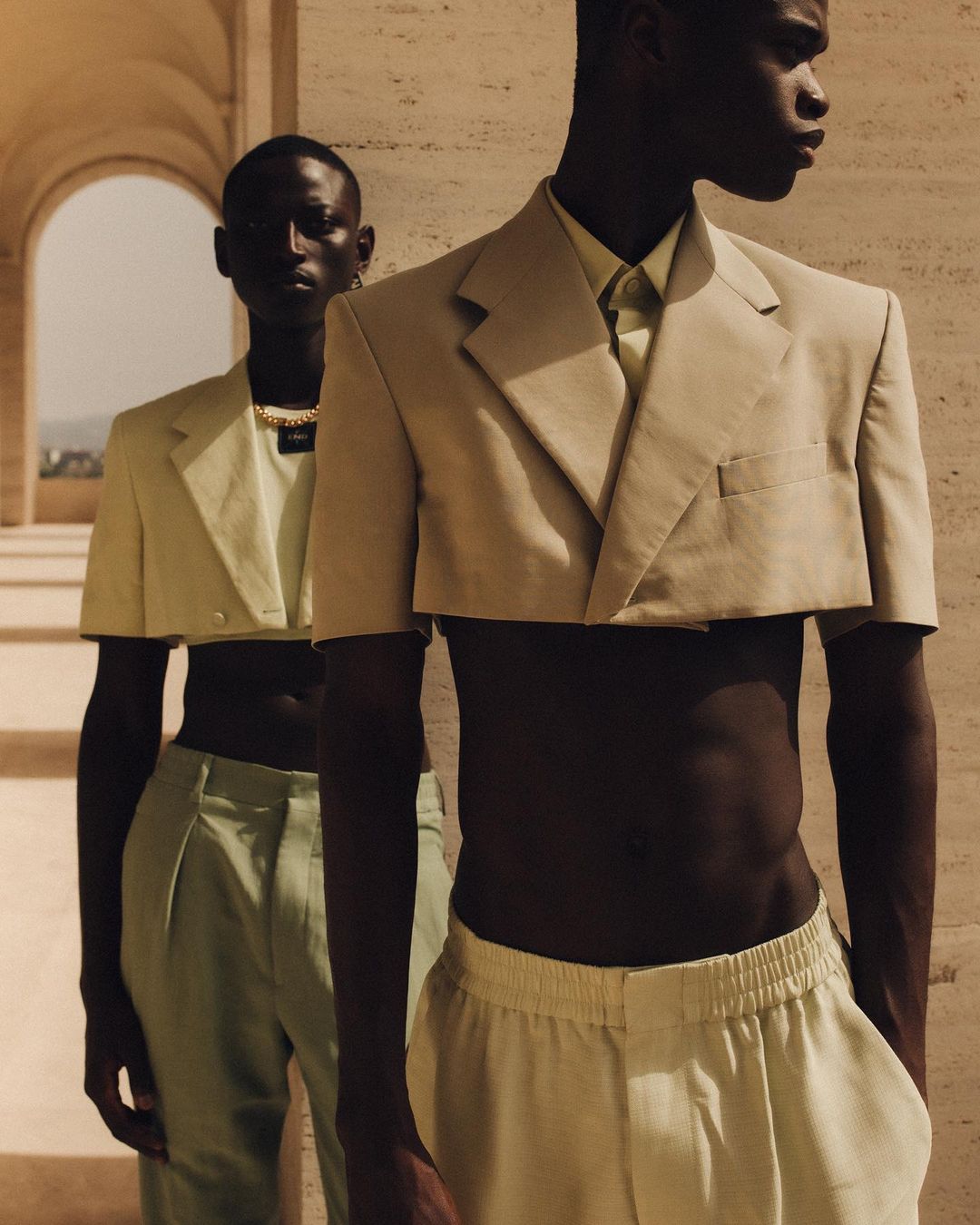 Two African male fashion models pose in cropped blazers and pants in front of a travertine colonnade, showcasing modern menswear.