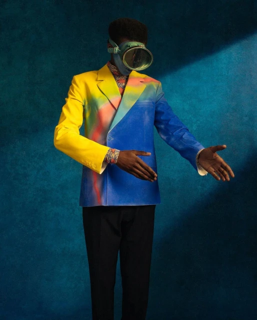 Fashion portrait of a man wearing a yellow and blue suit and a scuba mask standing against a blue backdrop with studio lighting.