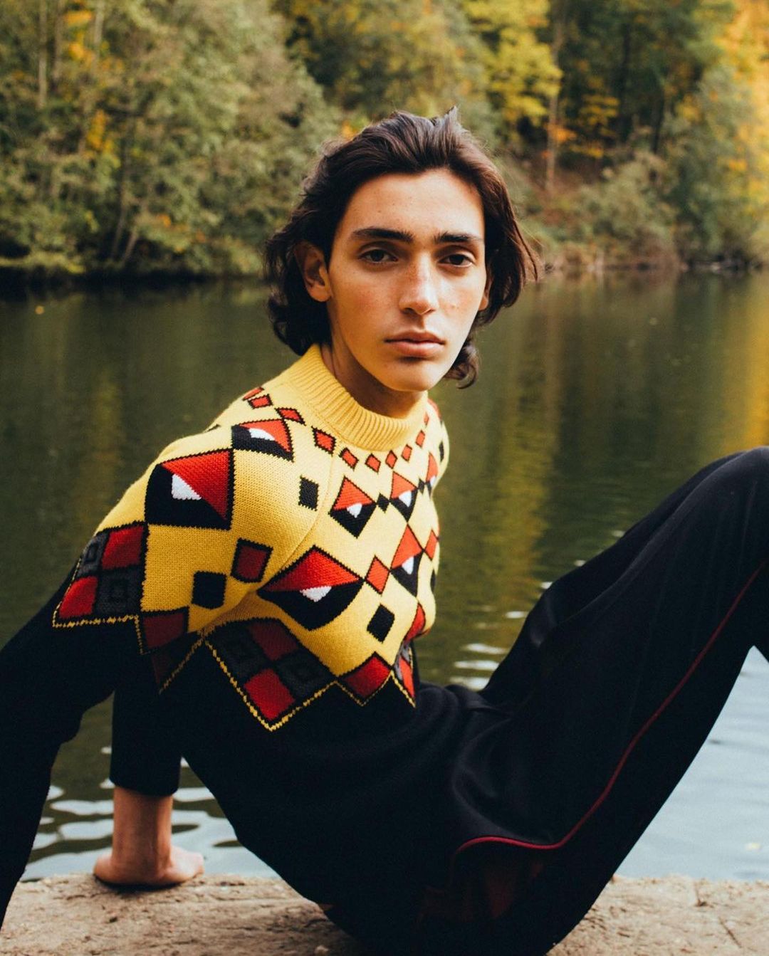 Young man with dark hair wearing a patterned yellow knit sweater by a lake, photographed by Antoine Henault