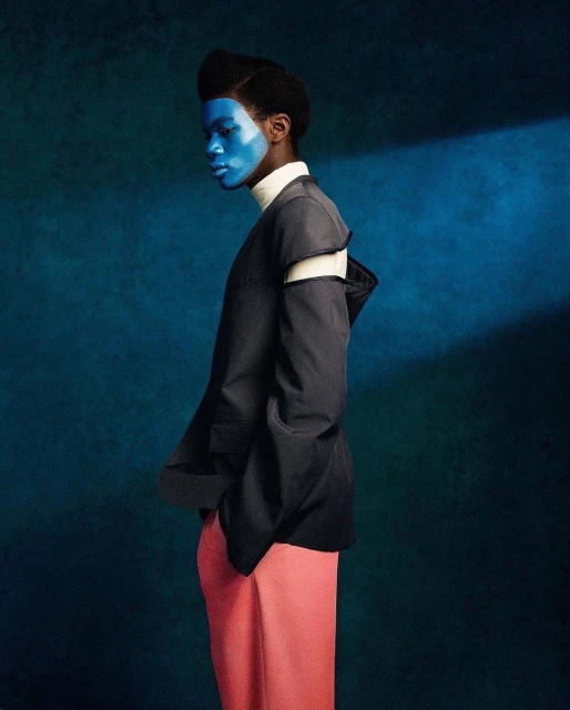 Side profile of a model with blue face paint wearing a dark grey jacket, white turtleneck, and pink pants against a dark blue background.