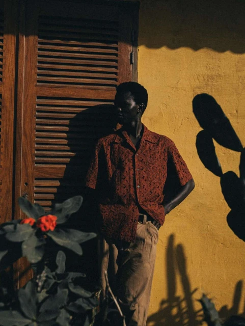 Stylish adult male in patterned shirt and khaki pants poses against yellow wall with wooden shutters and lush green plants.
