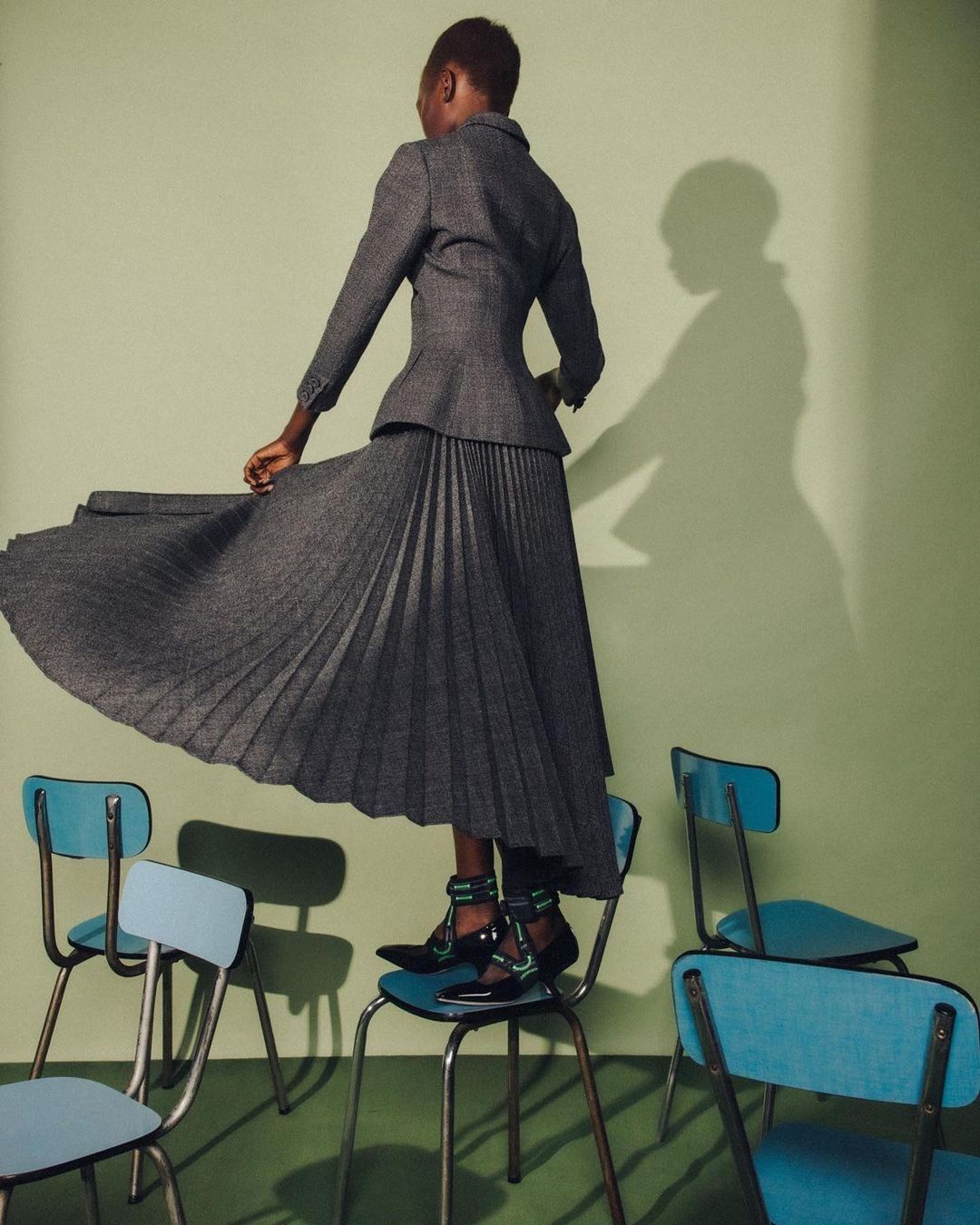 Woman in a gray suit with a pleated skirt poses on top of a stack of blue chairs in a fashion shot against a light green wall.
