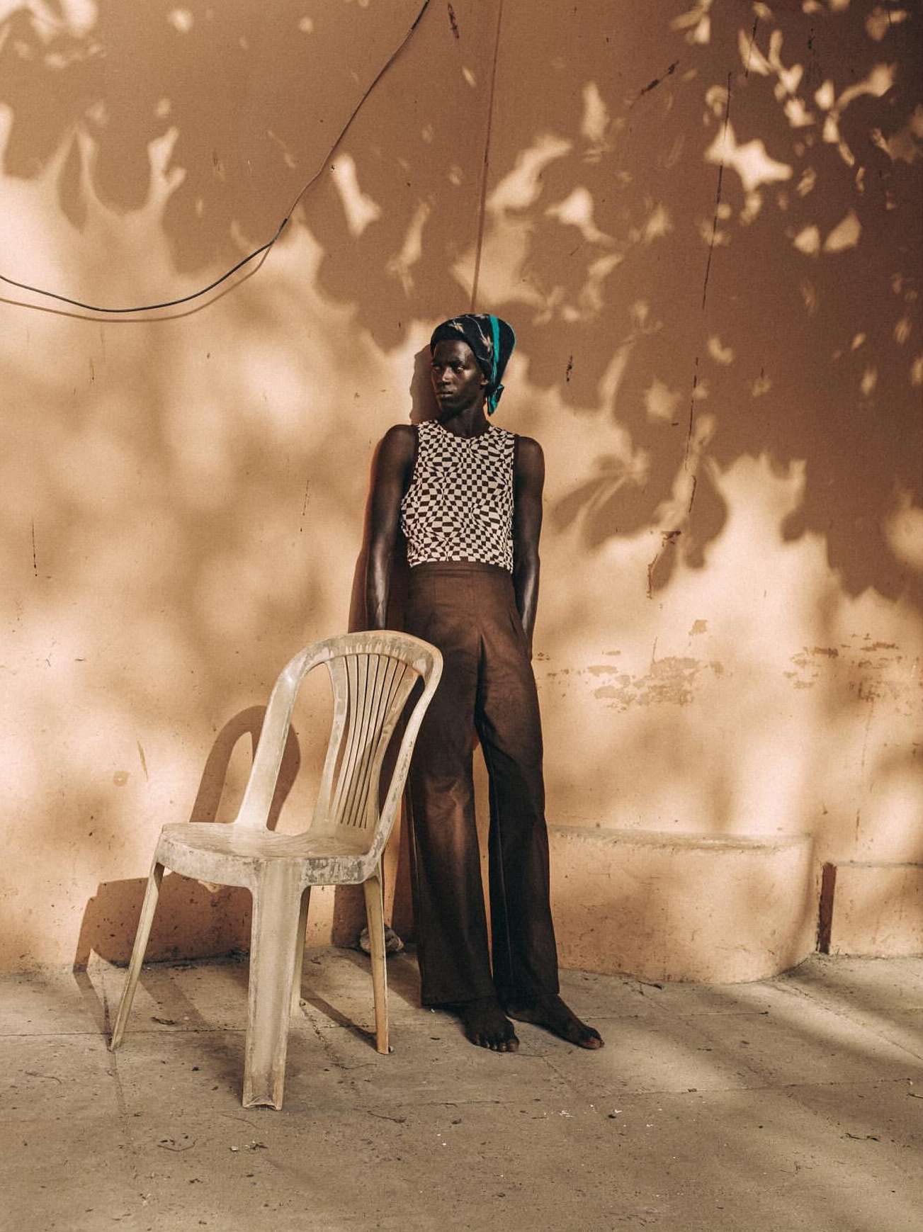 Portrait of an African woman in a patterned top and headscarf standing in front of a peach-colored wall with shadows, next to a white plastic chair.