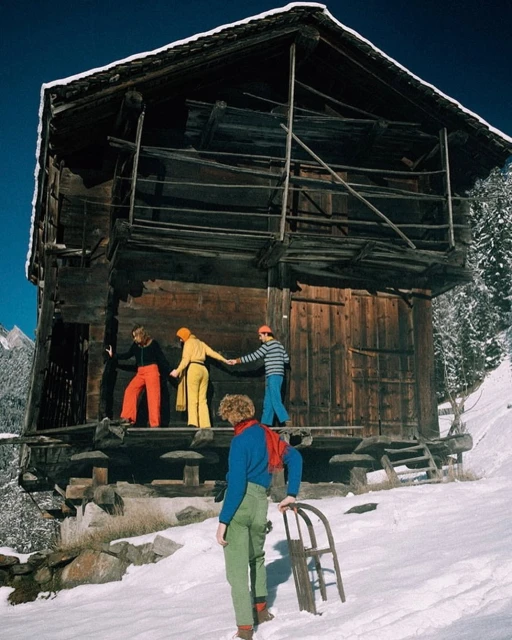 Four people in brightly colored outfits stand outside a weathered wooden building in a snowy, mountainous landscape with a vintage aesthetic.