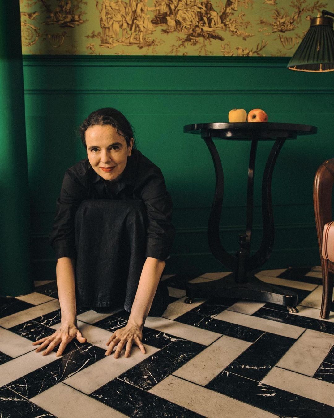 Portrait of a woman crouching in a dark dress on a black and white tiled floor with a dark table and green wall in the background.