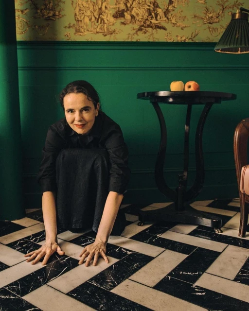 Portrait of a woman crouching in a dark dress on a black and white tiled floor with a dark table and green wall in the background.