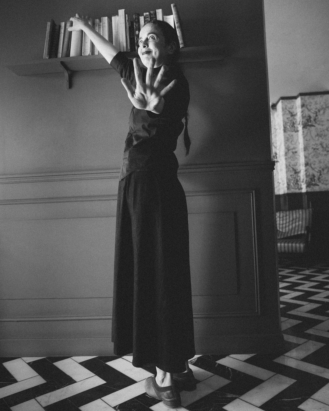 Monochrome shot of an adult female with braided hair, wearing a black outfit, standing near a shelf of books with her hand raised.