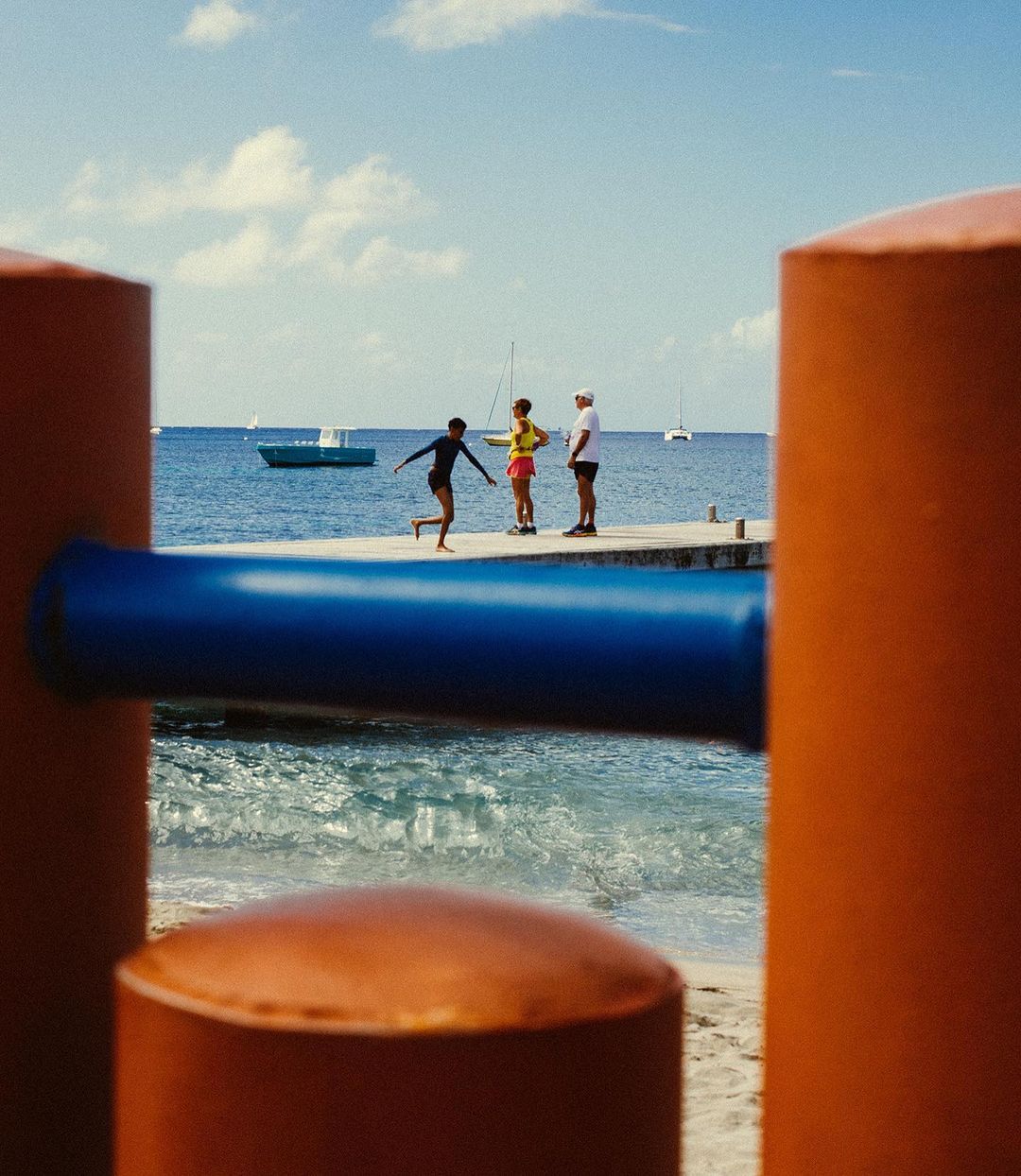 People on a pier near the ocean with boats in the distance and bright, colorful structures in the foreground on a sunny day.