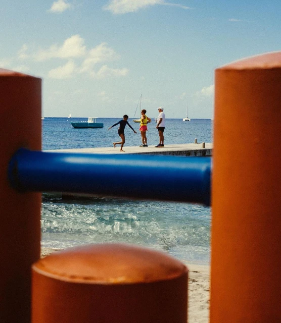 People on a pier near the ocean with boats in the distance and bright, colorful structures in the foreground on a sunny day.