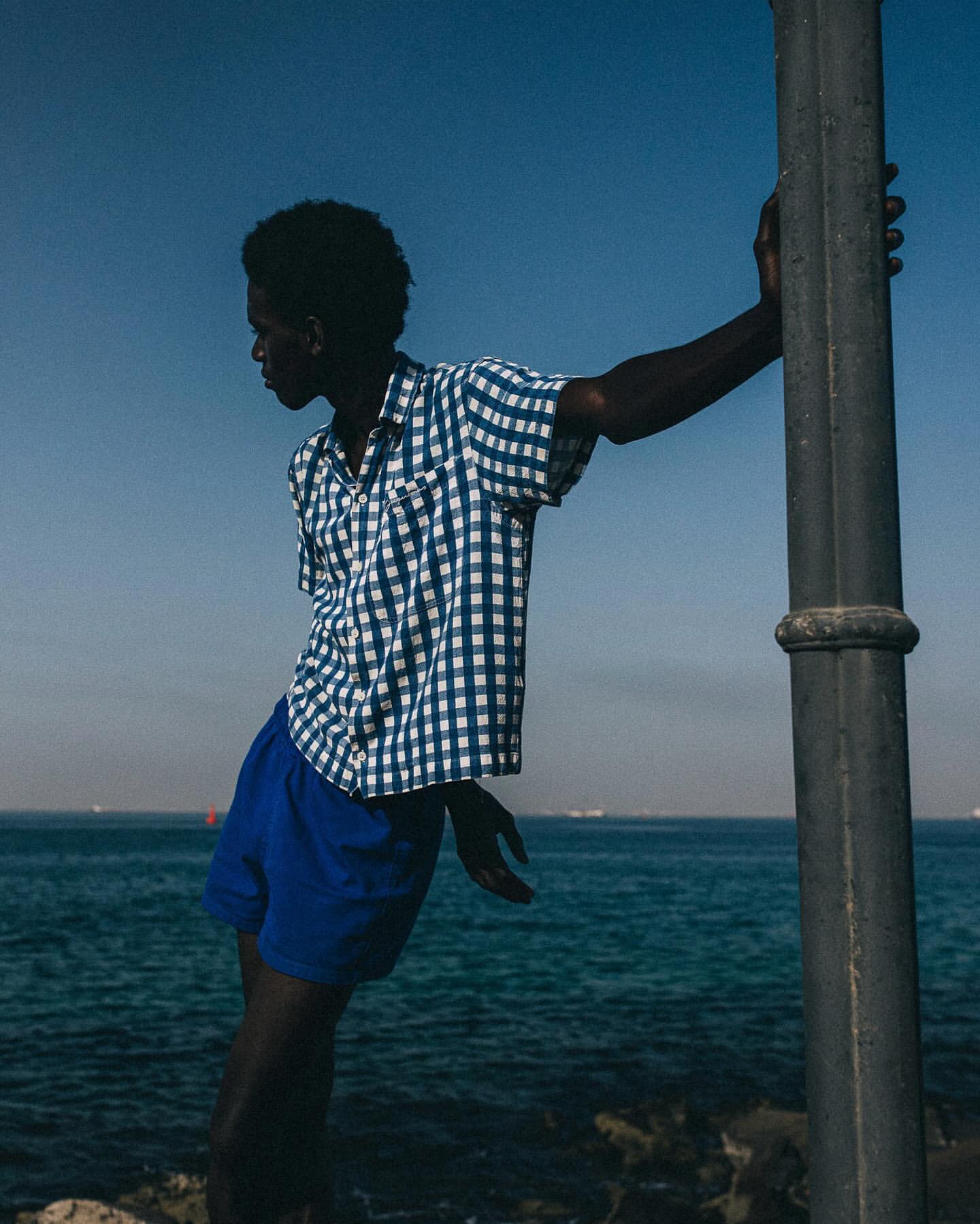 Young man poses near the ocean, wearing a blue and white checkered short-sleeve shirt and blue shorts, leaning against a grey pole.