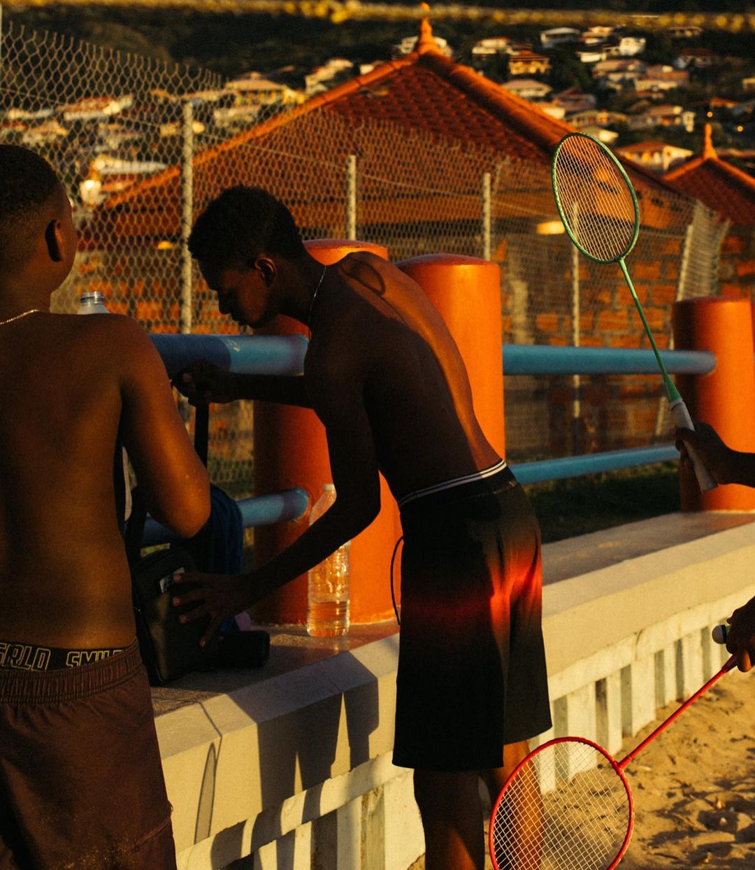 Outdoor recreation: Two young Black men are playing badminton in bright sunlight, with residential buildings and a chain-link fence behind them.