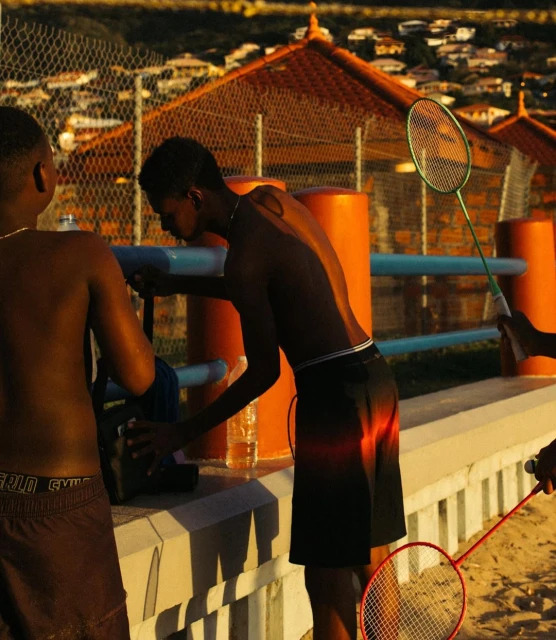 Outdoor recreation: Two young Black men are playing badminton in bright sunlight, with residential buildings and a chain-link fence behind them.