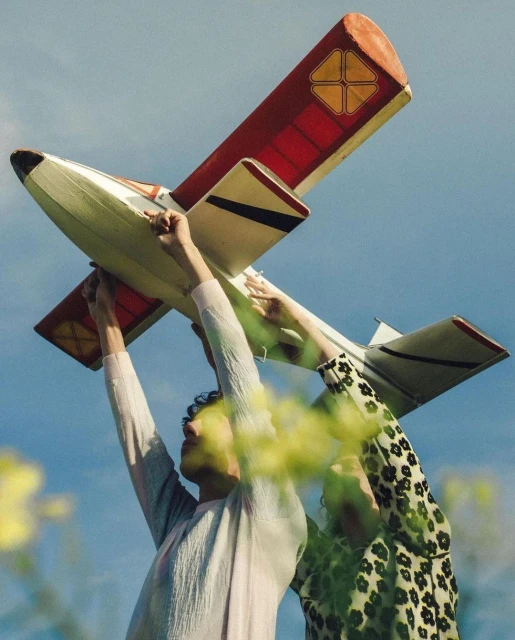 Two people holding a vintage model airplane up to the sky, shot at a low angle with visible wildflowers and clouds.