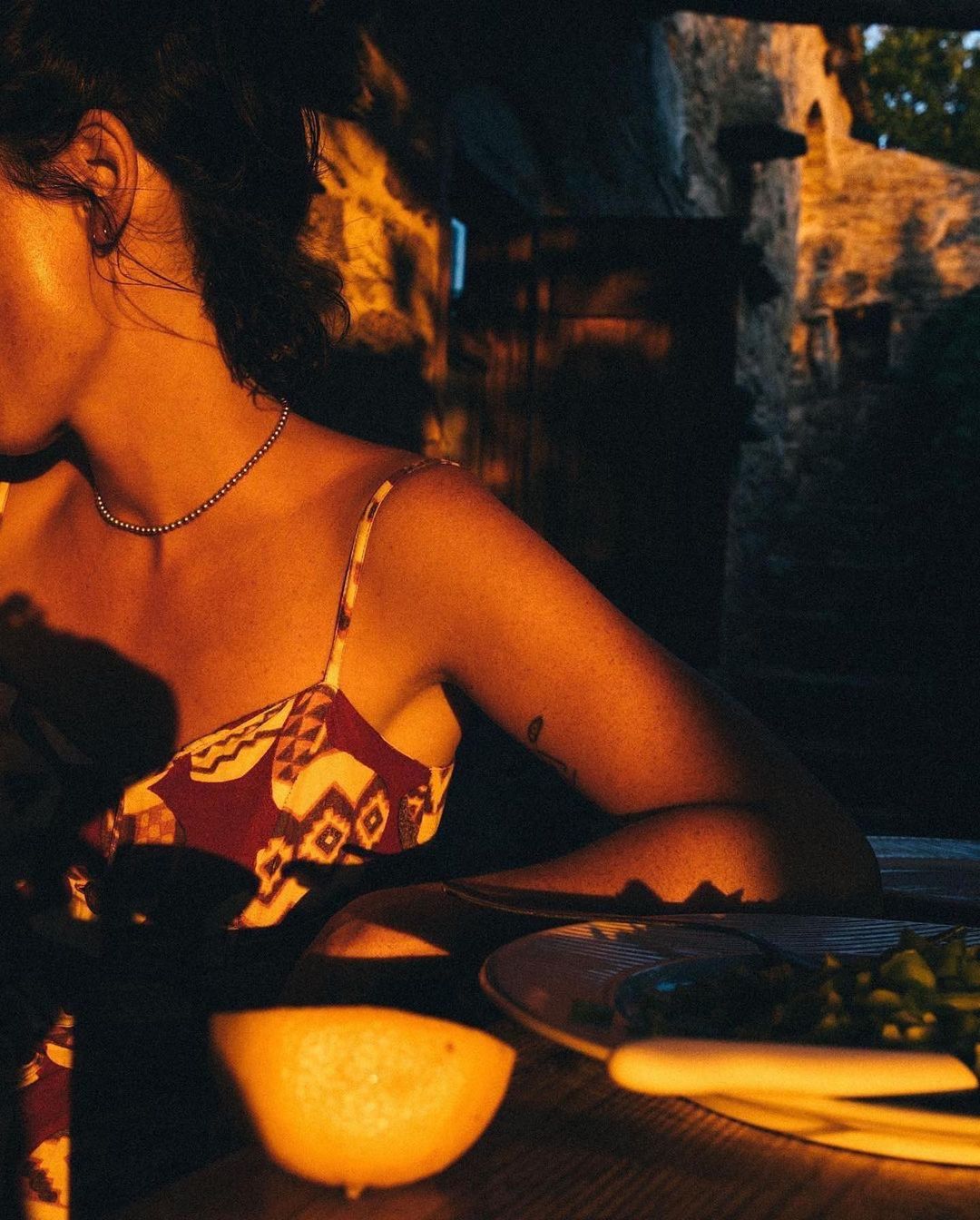 Woman in patterned dress sits at table with lemon and plate, bathed in warm, low light near stone wall in a dark, intimate setting.