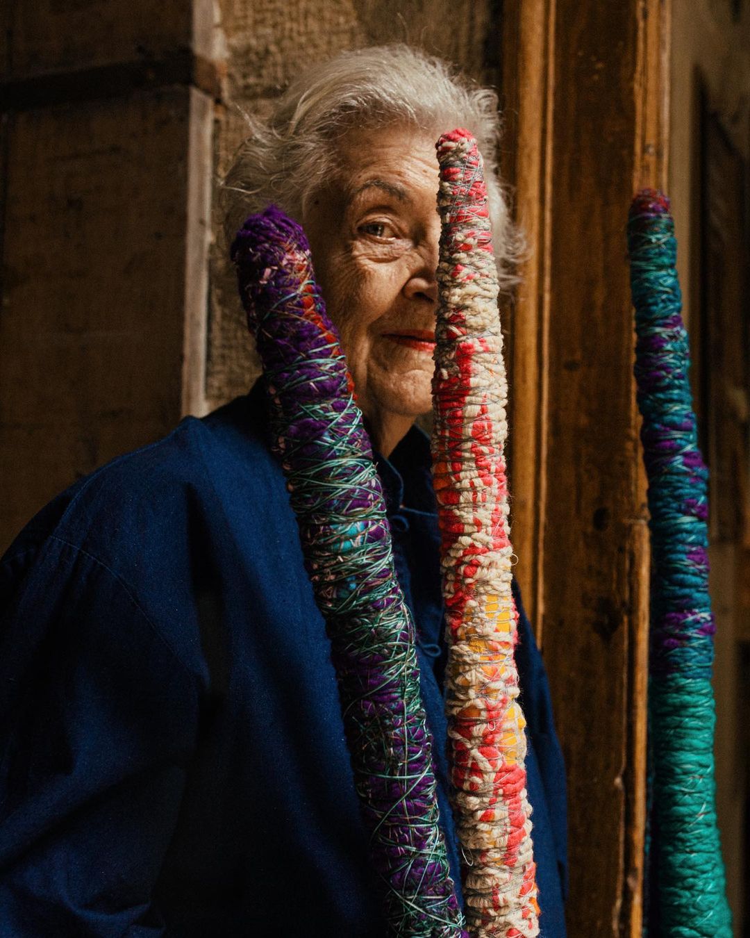 Elderly woman poses with colorful crafted yarn posts in an indoor setting with natural light, wearing a blue jacket.