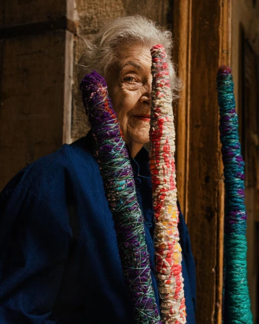 Elderly woman poses with colorful crafted yarn posts in an indoor setting with natural light, wearing a blue jacket.
