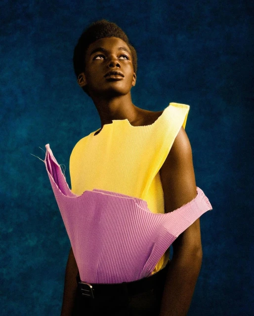 Portrait of a young Black male model wearing an open yellow pleated top over a pink top against a dark blue textured backdrop.
