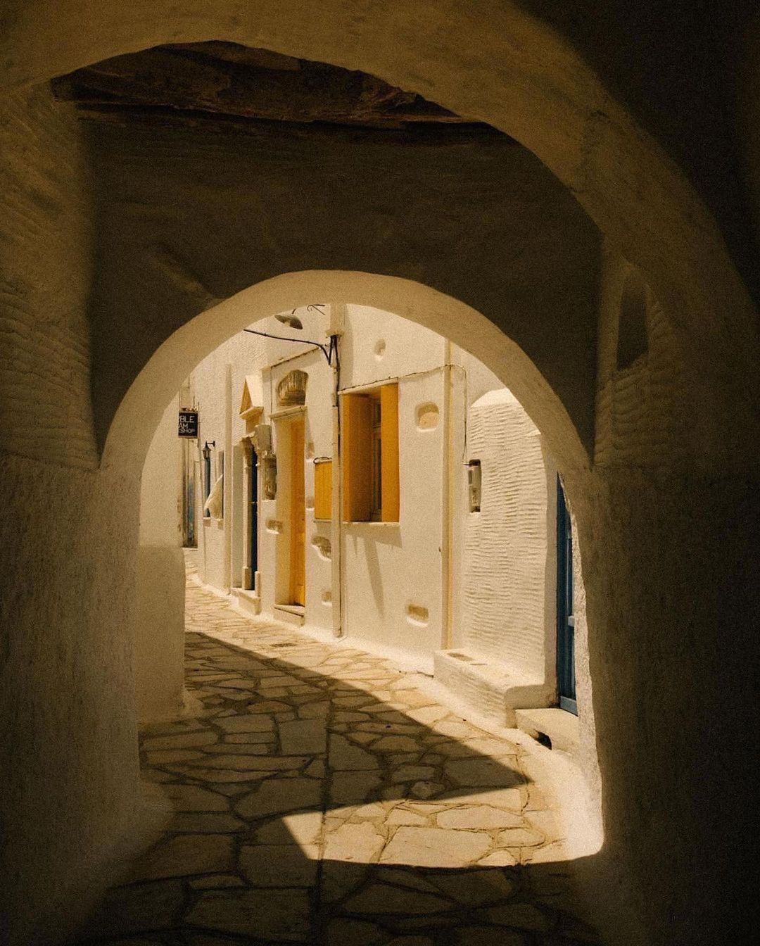 Archway frames a cobblestone alley in a Greek village, showcasing traditional white buildings and shadows in a warm, inviting scene.