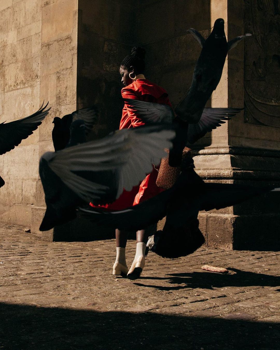 Woman in red jacket stands among pigeons in an urban area with sunlit stone architecture and cobblestone ground.
